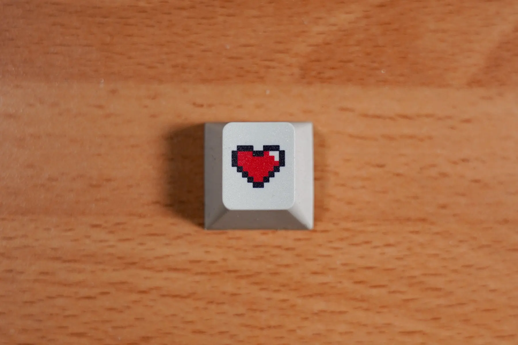A single white keyboard key printed with a small red pixel heart, resting on a wooden desk